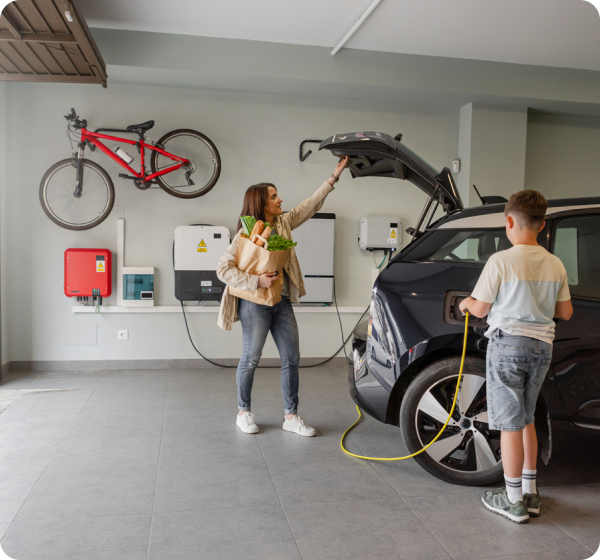 A mom in her garage taking groceries out of the back of her EV while her son plugs the charger in